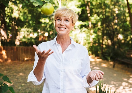 Woman in white collared shirt smiling with throwing green apple outside