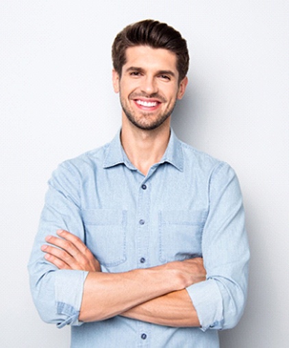 A young man wearing a blue button-down shirt with his arms crossed, smiling after having metal-free dentistry in Saginaw to restore his smile